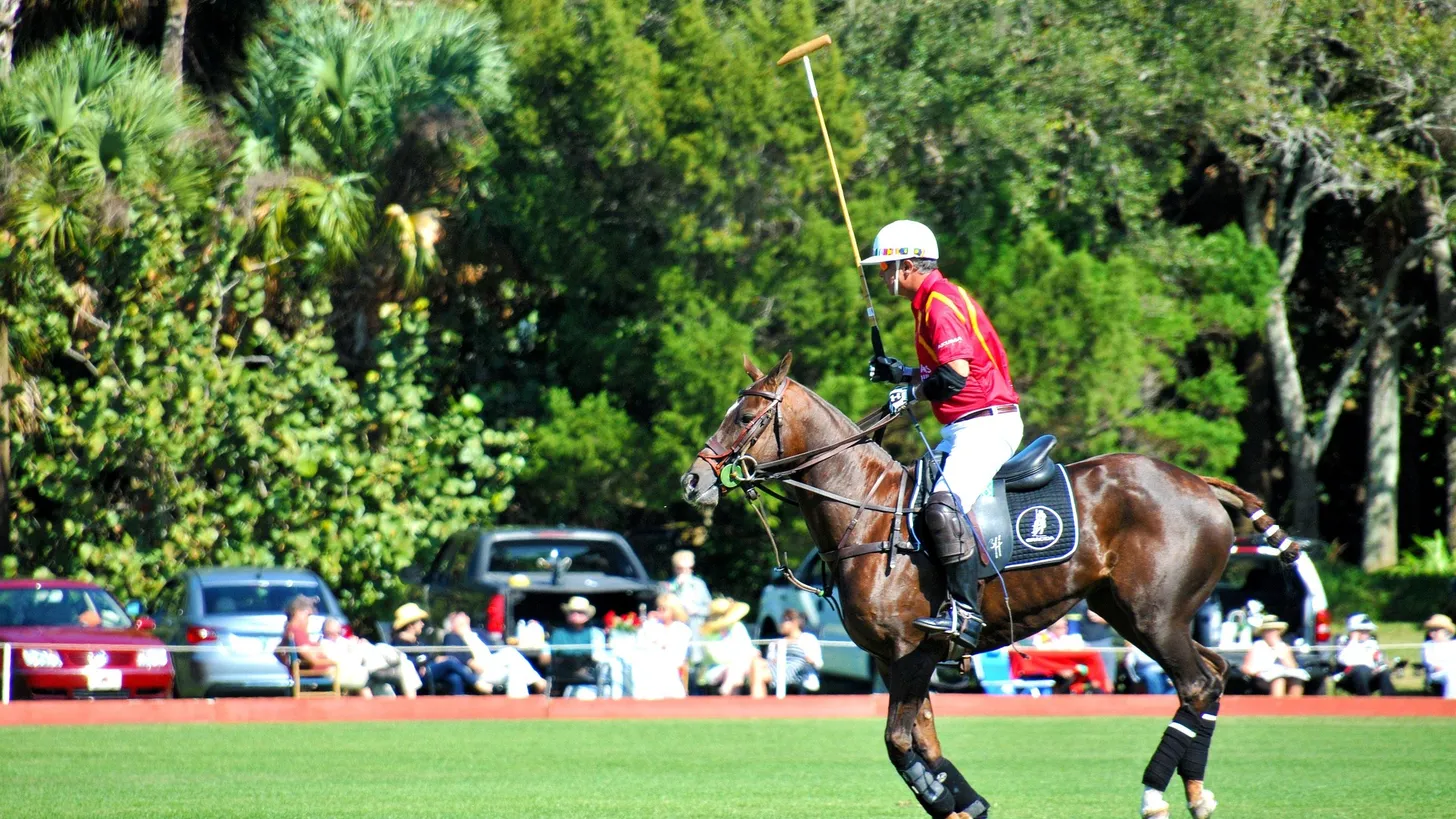 Jugador de polo con chomba roja galopando con el taco en alto durante un partido, con el público sentado al costado de la cancha
