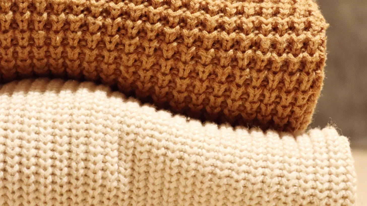 Pila de chombas de lana dobladas en tonos crema, camel y beige, con distintos tipos de punto a la vista