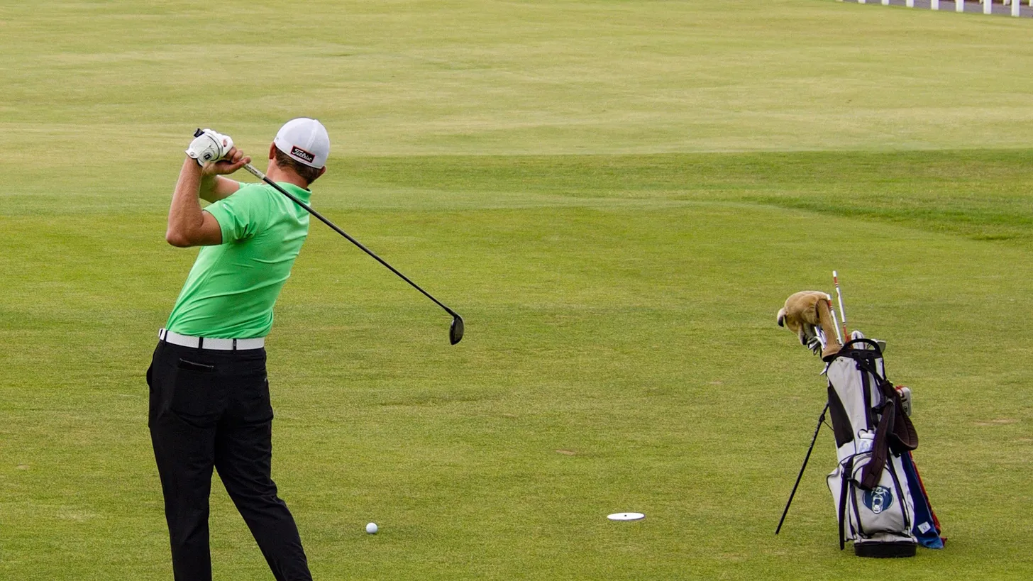 Golfista de chomba verde claro terminando el swing en el fairway de una cancha de links, con la bolsa de palos apoyada a un costado